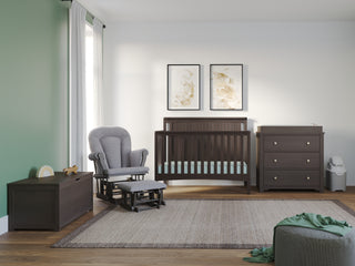 A modern nursery features a dark wood crib, matching dresser, gray glider with ottoman, Child Craft Harmony 33" Kids Toy Box in Dapper Gray, soft rug, green pouf, two framed art prints on white and green walls, and natural light from a window.