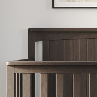 A close-up of the Child Craft Scout 4-in-1 Convertible Crib in Dapper Gray, featuring vertical slats against a light wall. The edge of a framed picture above adds extra charm to the baby nursery.