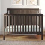 The Child Craft Scout 4-in-1 Convertible Crib in Dapper Gray features vertical slats and a beadboard headboard, holds a light mattress, and stands on a wooden floor with two framed botanical prints displayed behind it.