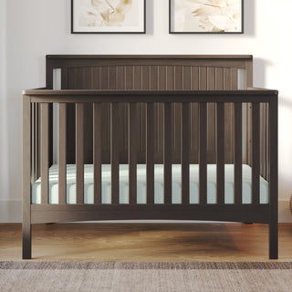 The Child Craft Scout 4-in-1 Convertible Crib in Dapper Gray features vertical slats and a beadboard headboard, holds a light mattress, and stands on a wooden floor with two framed botanical prints displayed behind it.