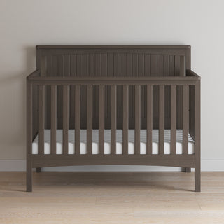 The Child Craft Scout 4-in-1 Convertible Crib in Dapper Gray features vertical slats and a simple headboard, includes a white mattress, and stands on a light wood floor against a plain light gray wall—ideal nursery furniture.