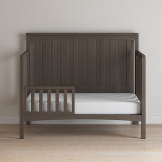 The Child Craft Scout 4-in-1 Convertible Crib in Dapper Gray features a slatted headboard, a partial left side rail, and includes a white mattress. It is shown against a light wall on a wooden floor.