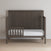The Child Craft Scout 4-in-1 Convertible Crib in Dapper Gray features a slatted headboard, a partial left side rail, and includes a white mattress. It is shown against a light wall on a wooden floor.
