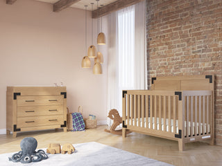 Modern nursery with a wooden crib and Child Craft’s Lucas Dresser in Nutmeg, both with black metal accents. Pendant lights, a brick accent wall, toys, and a woven basket sit near a window with sheer curtains.
