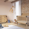 A modern nursery with a wooden crib, matching dresser featuring the Child Craft F01792 Universal Changing Table Topper in Nutmeg, pendant lamps for soft lighting, a plush rug, toys on the floor, wicker basket, rocking horse, and a brick accent wall.