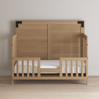 The Lucas 4-in-1 Convertible Crib by Child Craft in Nutmeg features slatted sides, a partial safety rail, and a tall paneled headboard, set against a light wall on a light wood floor.