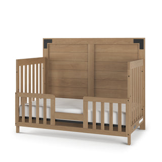 The Child Craft Lucas 4-in-1 Convertible Crib in Nutmeg features slatted sides, an open front, a paneled headboard with black corner accents, and a light brown finish. A white mattress rests inside the frame.
