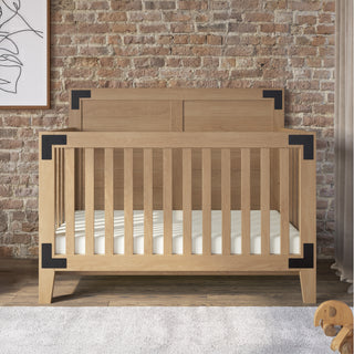 The Child Craft Lucas 4-in-1 Convertible Crib in Nutmeg, featuring black metal corner accents and vertical slats with a white mattress, stands by a brick wall with abstract art above and a plush dog toy on the soft rug nearby.