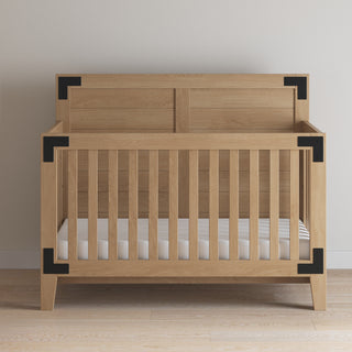 The Child Craft Lucas 4-in-1 Convertible Crib in Nutmeg features vertical slats and black metal corner brackets, and is shown on a light wood floor against a plain white wall.