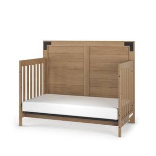 The Child Craft Lucas 4-in-1 Convertible Crib in Nutmeg features a high slatted back, partial side rails, a white mattress, and simple modern design with black metal corner accents.