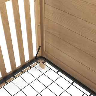 Close-up of the Child Craft Lucas 4-in-1 Convertible Crib in Nutmeg, highlighting its wooden slatted and panel sides, metal mattress support frame, and a small wooden support block underneath.