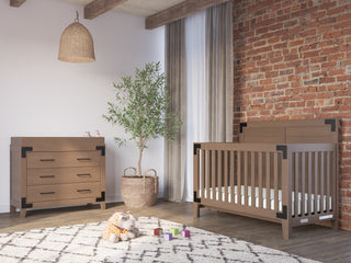 A cozy nursery features the Child Craft Lucas 4-in-1 Convertible Crib in Dusty Heather, a matching dresser, woven basket with plant, wicker lamp, plush toy and blocks on a rug, brick accent wall, and gray curtains over the window.