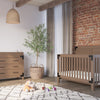 A cozy nursery with a wooden crib and matching dresser featuring the Child Craft F01792 Universal Changing Table Topper in Dusty Heather, a woven basket with a green plant, hanging light, plush toys on a rug, and sheer curtains by the window.