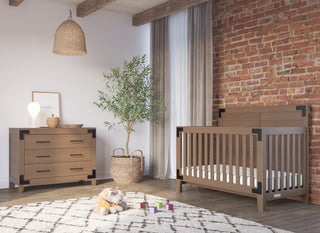 A cozy nursery features the Child Craft Lucas 4-in-1 Convertible Crib in Dusty Heather, a matching dresser, stuffed giraffe toy, colorful blocks, wicker basket with potted plant, exposed brick wall, wooden beams, and sheer curtains letting in light.