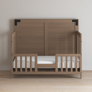 The Child Craft F09514 Toddler Guard Rail in Dusty Heather features sturdy wood construction with slatted sides and an open front, a high back panel, and is displayed against a plain light wall on a light wood floor.
