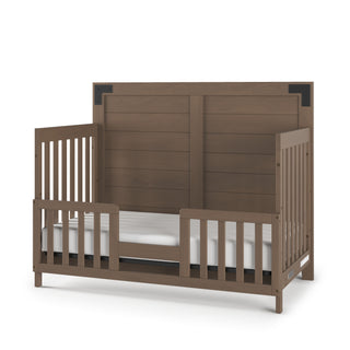 The Child Craft Lucas 4-in-1 Convertible Crib in Dusty Heather features a high headboard, safety rails, and a white mattress. It has a rustic brown finish with metal corner accents on the headboard.