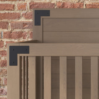 Close-up of the Child Craft Lucas 4-in-1 Convertible Crib in Dusty Heather, featuring vertical wooden slats and black metal corner brackets, set against a textured red brick wall.