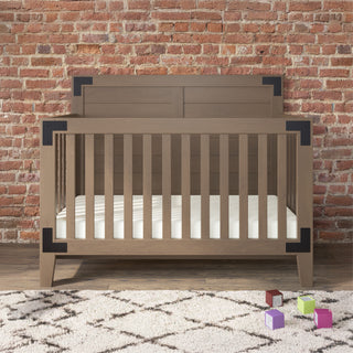 The Child Craft Lucas 4-in-1 Convertible Crib in Dusty Heather, featuring black metal corner accents, stands on a patterned rug before a brick wall, with four colorful toy blocks scattered nearby.