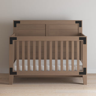 The Child Craft Lucas 4-in-1 Convertible Crib in Dusty Heather, featuring vertical slats and black metal corner brackets, is set against a plain light wall on a hardwood floor and comes with a white mattress.