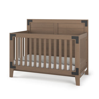 The Child Craft Lucas 4-in-1 Convertible Crib in Dusty Heather features vertical slats, dark metal corner brackets, a solid panel headboard, and comes with a white mattress inside.