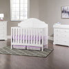 A bright nursery features a white crib, purple bedding, two dressers with the Child Craft F01792 Universal Changing Table Topper in matte white, a gray patterned rug, lamp, framed art, sunlit window, beige walls, and dark wood floors for warmth.