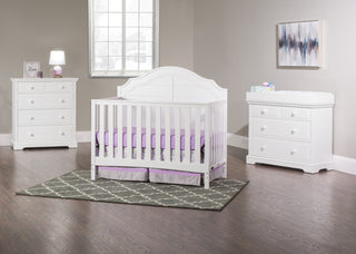 A bright nursery features a white crib, purple bedding, two dressers with the Child Craft F01792 Universal Changing Table Topper in matte white, a gray patterned rug, lamp, framed art, sunlit window, beige walls, and dark wood floors for warmth.