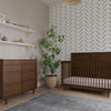 A modern nursery features a wooden crib, Child Craft dresser with the F01792 Universal Dressing Kit in Toasted Chestnut, a potted plant, two decor shelves, a chevron accent wall, and a gray rug on light wood floors.