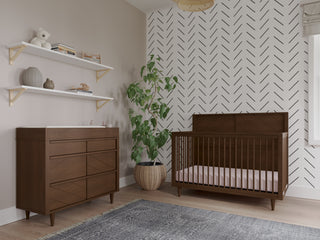 A modern nursery features a wooden crib, Child Craft dresser with the F01792 Universal Dressing Kit in Toasted Chestnut, a potted plant, two decor shelves, a chevron accent wall, and a gray rug on light wood floors.