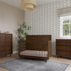 A modern nursery with brown wooden furniture by Child Craft, featuring the F01792 Universal Dressing Kit in Toasted Chestnut, a crib, dressers with changing topper, gray rug, potted plant, black chevron walls, and white-curtained window.