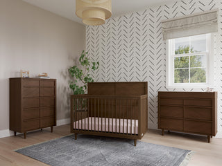 Modern nursery with a brown crib, Surrey Hill 3 Drawer Dresser by Child Craft in Toasted Chestnut, and chest of drawers on light wood floors. Features a plant, gray rug, geometric accent wall, Roman shade, and woven ceiling light.