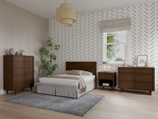 A modern bedroom with geometric wallpaper, a double bed with a brown headboard, two Child Craft Surrey Hill 3 Drawer Dressers in Toasted Chestnut, a nightstand, gray rug, Roman shade window, hanging light, and potted plant.
