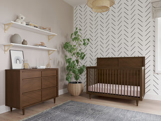 A modern nursery features a Child Craft Surrey Hill 3 Drawer Dresser in Toasted Chestnut, a wooden crib, potted plant, patterned accent wall, two white decor shelves, a gray rug, and light wood floors with soft natural light from a window.