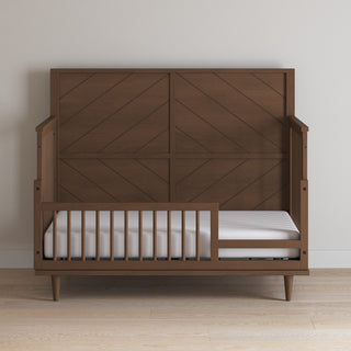 The Child Craft F09502 Toddler Guard Rail in Toasted Chestnut adds safety to a wooden toddler bed with a high paneled headboard and minimalist design, set on a light wood floor against a plain beige wall.