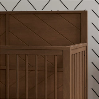 A close-up of the Child Craft Surrey Hill 4-in-1 Convertible Crib in Toasted Chestnut, featuring vertical slats and a diagonal panel design, stands against a white wall with a black, diagonal-line pattern.