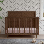 The Child Craft Surrey Hill 4-in-1 Convertible Crib in Toasted Chestnut with a pink mattress rests on a light wood floor. A black chevron pattern decorates the white wall, with wooden toys and a leafy plant nearby.