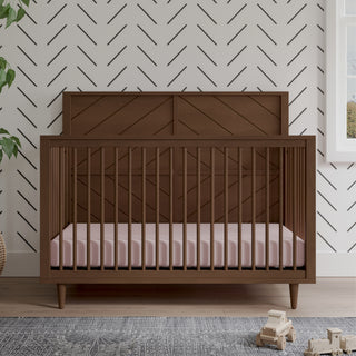 The Child Craft Surrey Hill 4-in-1 Convertible Crib in Toasted Chestnut with a pink mattress rests on a light wood floor. A black chevron pattern decorates the white wall, with wooden toys and a leafy plant nearby.