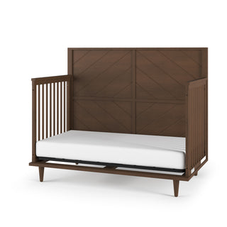 The Child Craft Surrey Hill 4-in-1 Convertible Crib in Toasted Chestnut features an open front, high solid chevron-patterned headboard, side railings, and a white mattress. The dark brown wood finish adds a stylish touch.