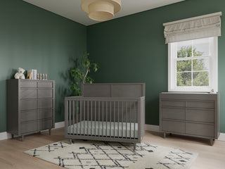 Modern nursery with dark green walls, a gray Child Craft Surrey Hill crib, Child Craft Surrey Hill 3 Drawer Dresser in Lunar Gray, gray chest, plush white rug with black diamond pattern, leafy plant, white blinds, and woven ceiling light.