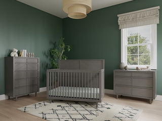 A modern nursery with green walls, a gray crib, a Child Craft Surrey Hill 3 Drawer Dresser in Lunar Gray, a matching chest, a white rug with a black geometric pattern, a potted plant, a roman shade, and a woven ceiling light.