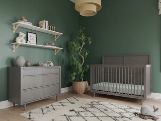A modern nursery with green walls, a gray crib, a Child Craft Surrey Hill 3 Drawer Dresser in Lunar Gray, potted plant, shelves with toys and books, patterned rug, woven ceiling light, and toy train and car on the wooden floor.