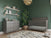 A modern nursery with green walls, a gray crib, a Child Craft Surrey Hill 3 Drawer Dresser in Lunar Gray, potted plant, shelves with toys and books, patterned rug, woven ceiling light, and toy train and car on the wooden floor.