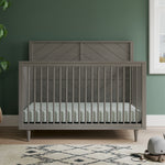 The Child Craft Surrey Hill 4-in-1 Convertible Crib in Lunar Gray, featuring a chevron-patterned headboard, stands against a green wall with part of a rug, a plant, and a framed picture also visible in the room.