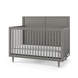 The Child Craft Surrey Hill 4-in-1 Convertible Crib in Lunar Gray features vertical slats on three sides, a chevron-patterned solid headboard, a white mattress, and stands on four angled legs.