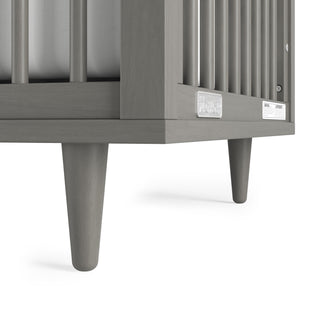 Close-up of the corner of a Child Craft Surrey Hill 4-in-1 Convertible Crib in Lunar Gray, featuring vertical bars, tapered legs, and a clear manufacturer label on the side.