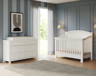 A modern nursery with blue striped and paneled walls, a white crib, the Child Craft Westgate Double Dresser in Vintage Linen with changing pad, wall lamps, a window with sheer curtains, and a neutral rug on a wooden floor.