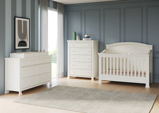 A modern nursery with white furniture, including a pine crib and the Child Craft Westgate 5-Drawer Chest in Vintage Linen, set against blue paneled walls. A beige rug covers the wooden floor by a large window with sheer curtains.