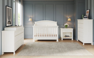 A modern nursery with gray paneled walls features the Child Craft Westgate 4-in-1 Convertible Crib in Vintage Linen, centered between wall sconces, with a white dresser, changing table, nightstand with flowers, and a light rug on hardwood floors.