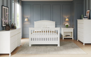 A spacious bedroom with blue paneled walls, a white crib on a gray rug, pine nursery dresser, the Child Craft Westgate 5-Drawer Chest in Vintage Linen, a white nightstand with a flower vase, modern wall lamps, and framed art on the walls.