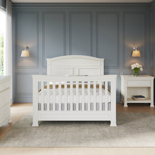 A stylish nursery features a white crib on a gray rug, flanked by matching Child Craft Westgate Nightstand in Vintage Linen, with blue paneled walls, wall sconces, and natural light streaming through the window.