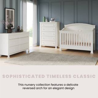 A stylish nursery with a white crib, Child Craft Westgate Double Dresser in Vintage Linen, and a chest of drawers against gray paneled walls, a large window, and a soft rug. “Sophisticated Timeless Classic – Delicate reversed arch for elegant design.”.